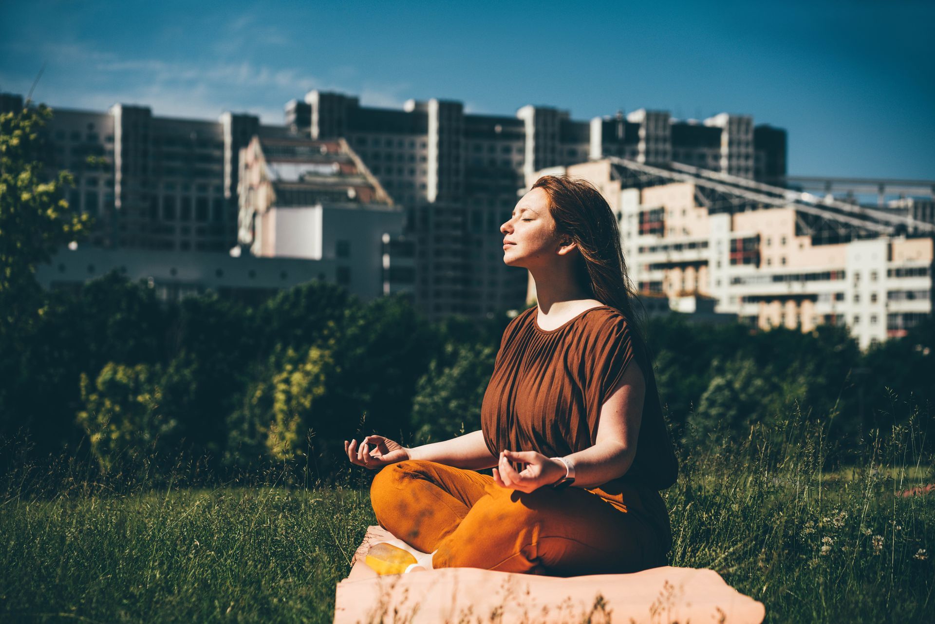 Woman meditating outdoors in a park, eyes closed, hands in prayer pose. Brown top, orange pants, city buildings in background.