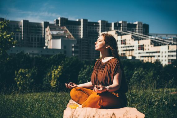 Woman meditating outdoors in a park, eyes closed, hands in prayer pose. Brown top, orange pants, city buildings in background.