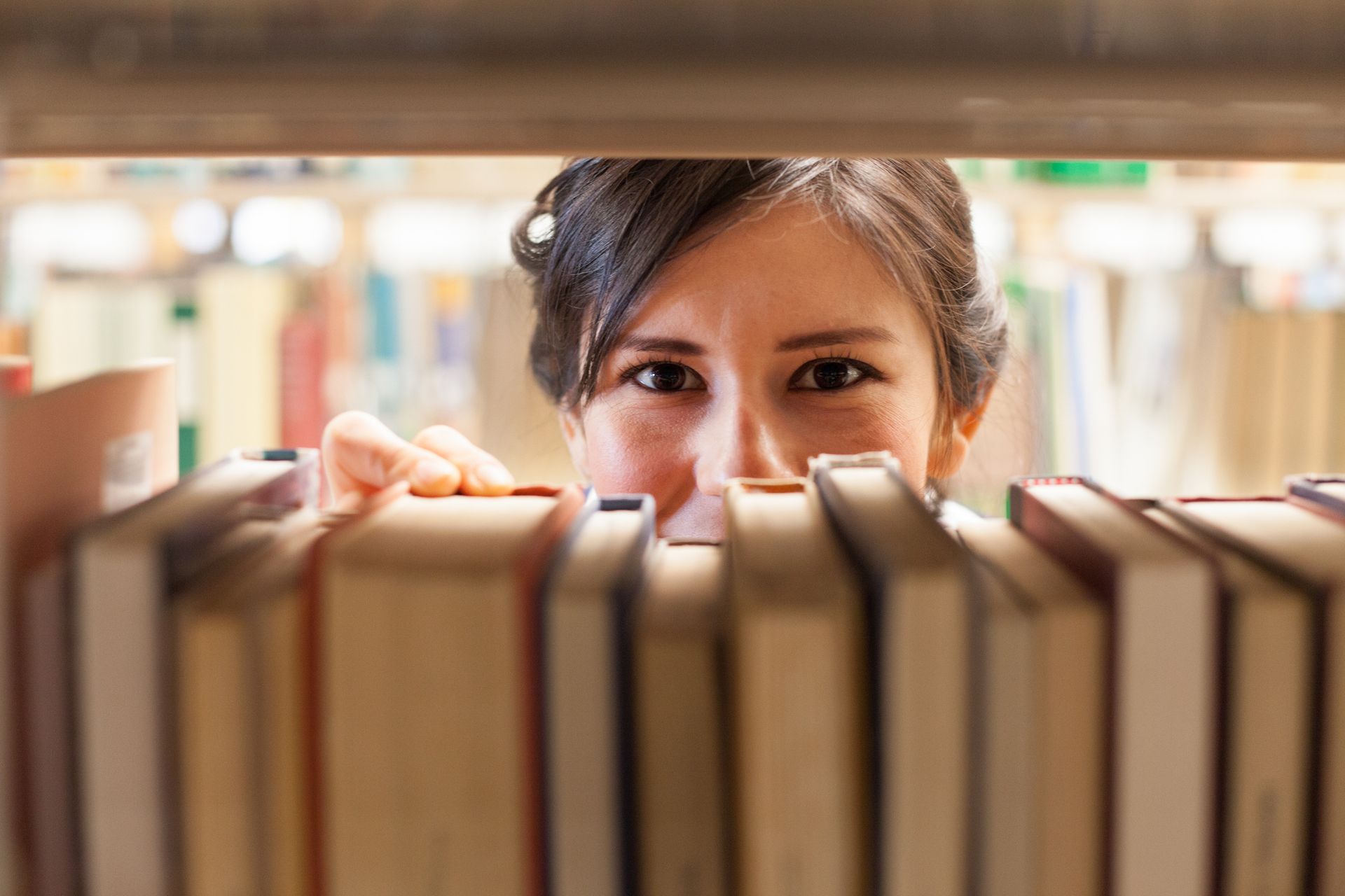 Woman peers out from behind a row of books on a library shelf, looking intently.