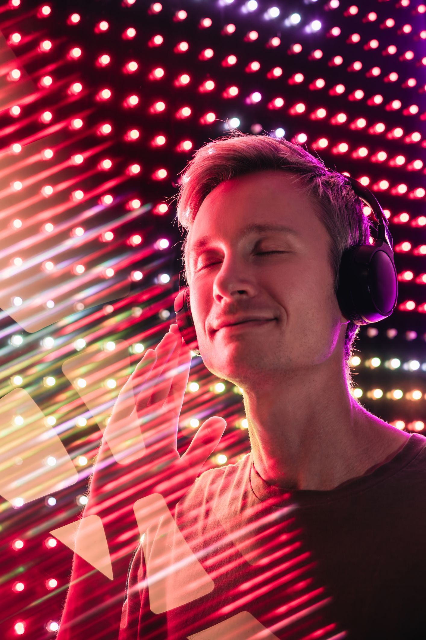 Man wearing headphones with eyes closed, enjoying music in front of a colorful, illuminated wall with red and white lights.