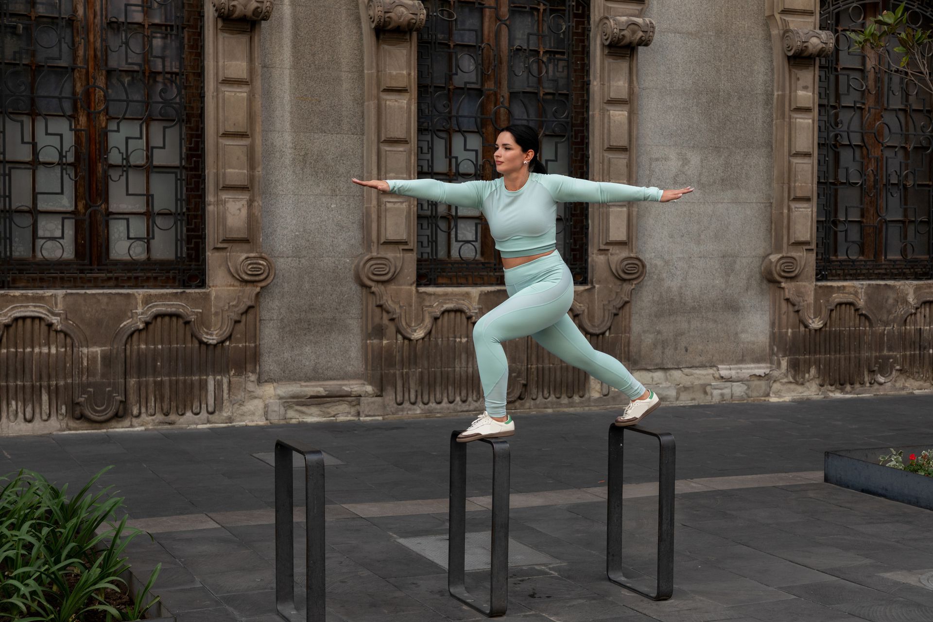 Woman in teal workout gear balances on three metal posts, arms outstretched, in front of a stone building.