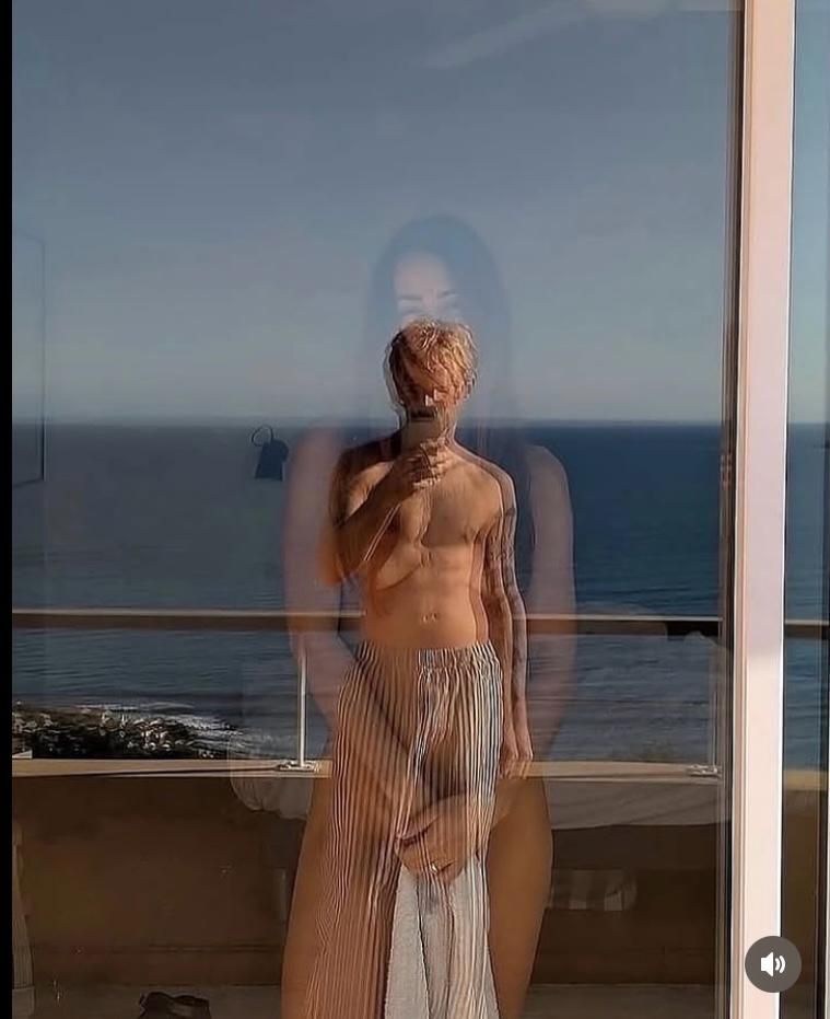 A shirtless person, possibly male, stands near a window. Their reflection is visible, with the ocean visible beyond the window.