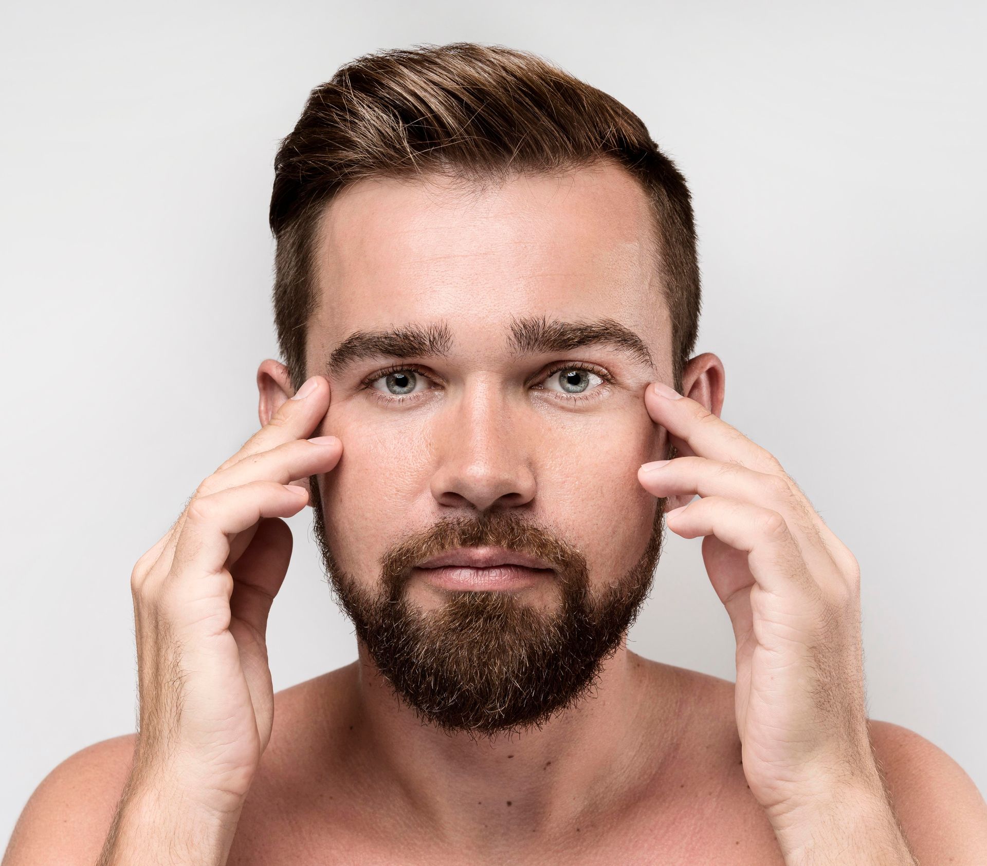 A shirtless man with a beard is touching his eyebrows with his hands.