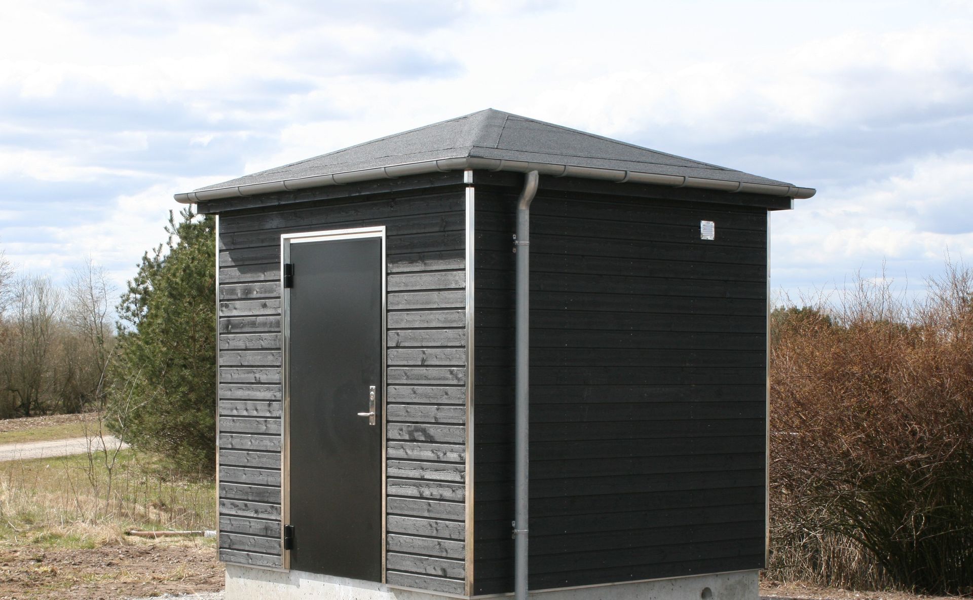 Et lille sort skur med sort dør står midt på en mark.
