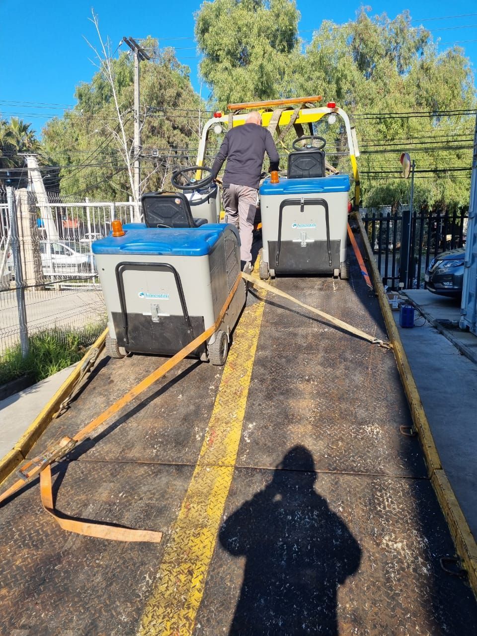 Persona cargando dos máquinas de limpieza de color gris/azul en un camión de plataforma con franjas amarillas.