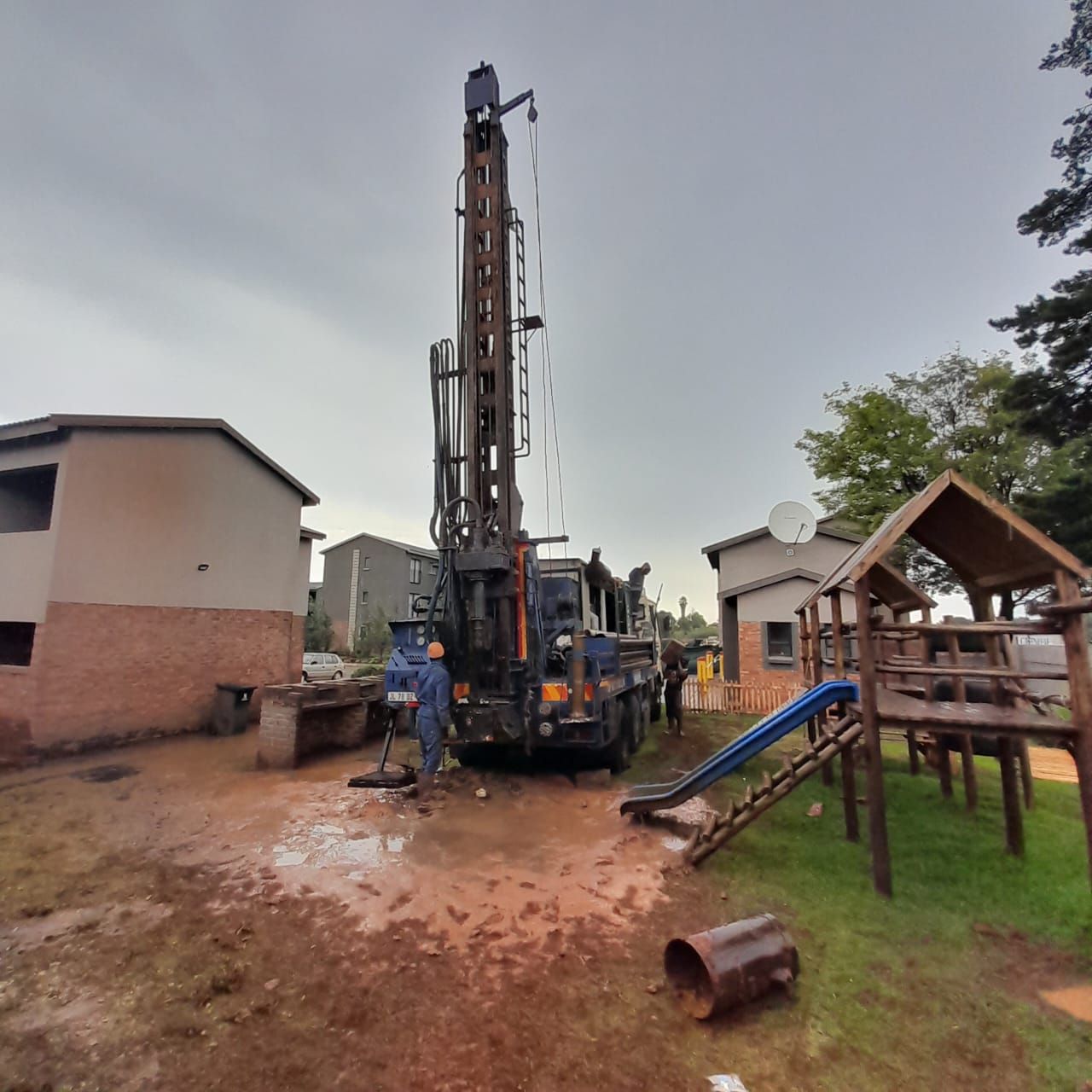 Borehole drilling machinery and workers on muddy field next to housing