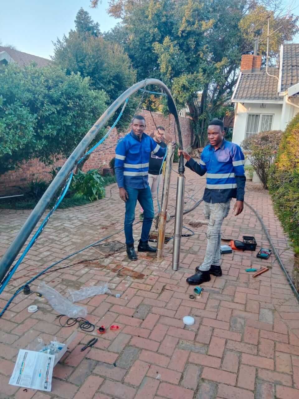 Two men are working on a pipe on a brick driveway.