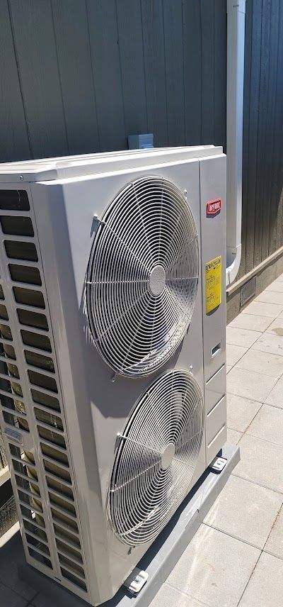 An air conditioning unit outdoors, with two fan grills and a white pipe next to a gray wall.