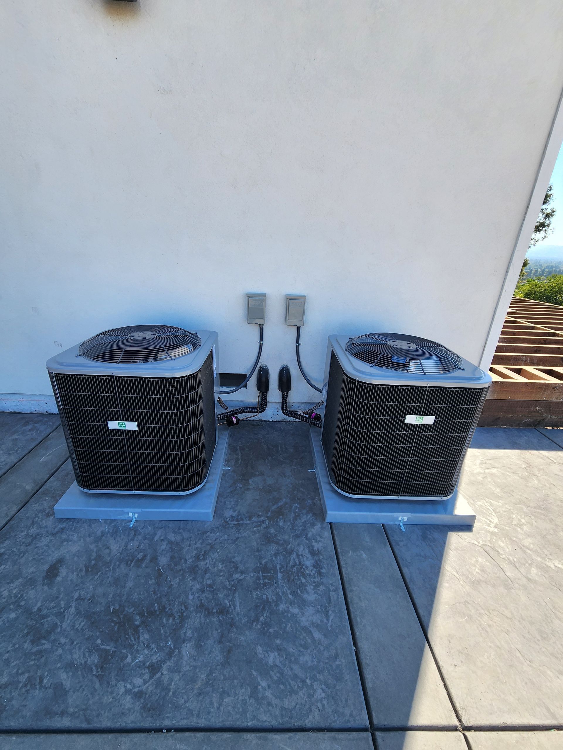 Two air conditioning units on a rooftop, with electrical conduit and a bright exterior wall.