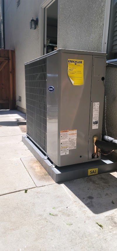 An air conditioning unit outdoors on a concrete pad. Grey unit with yellow sticker.
