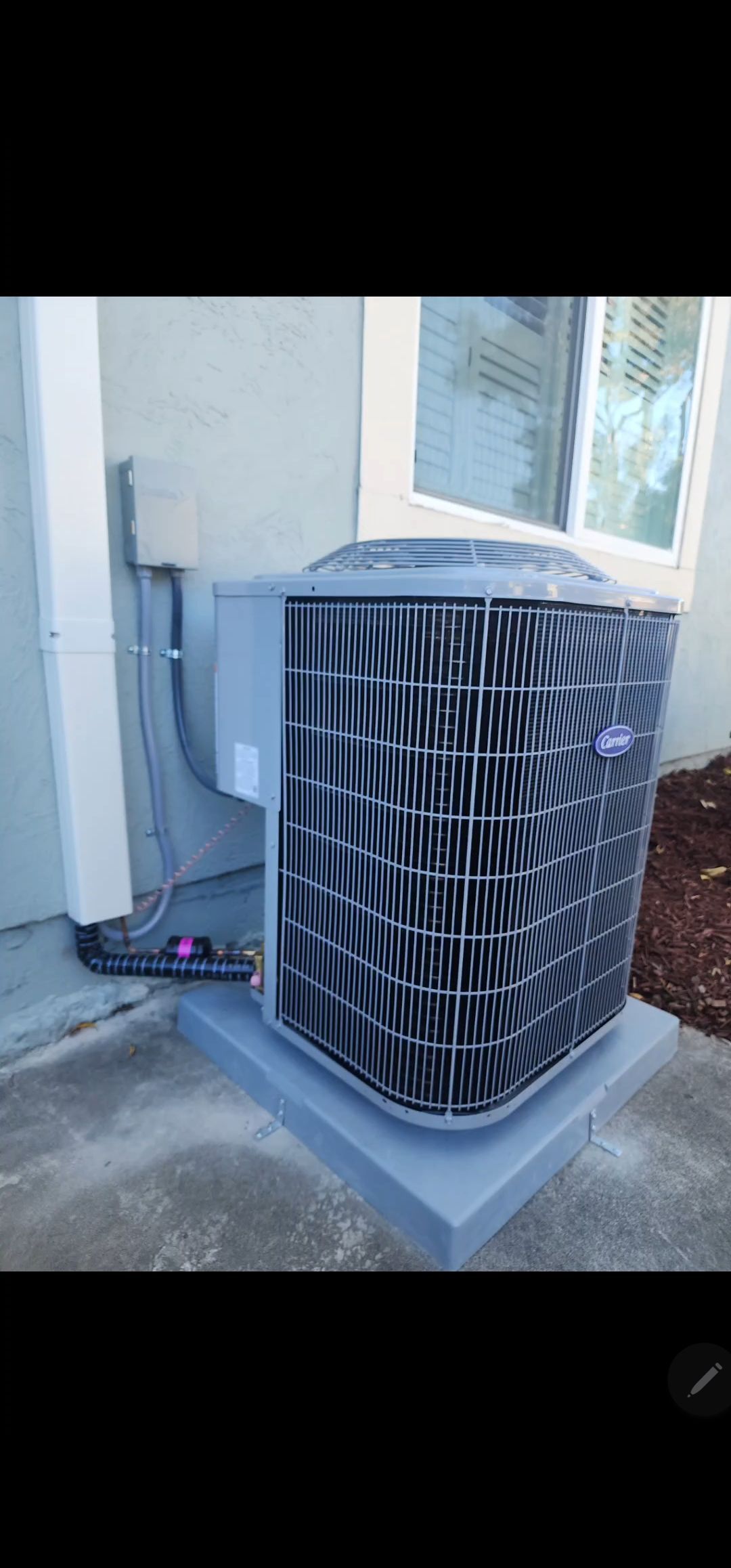 Outdoor air conditioning unit, gray and black, on a gray concrete pad, next to a building wall.