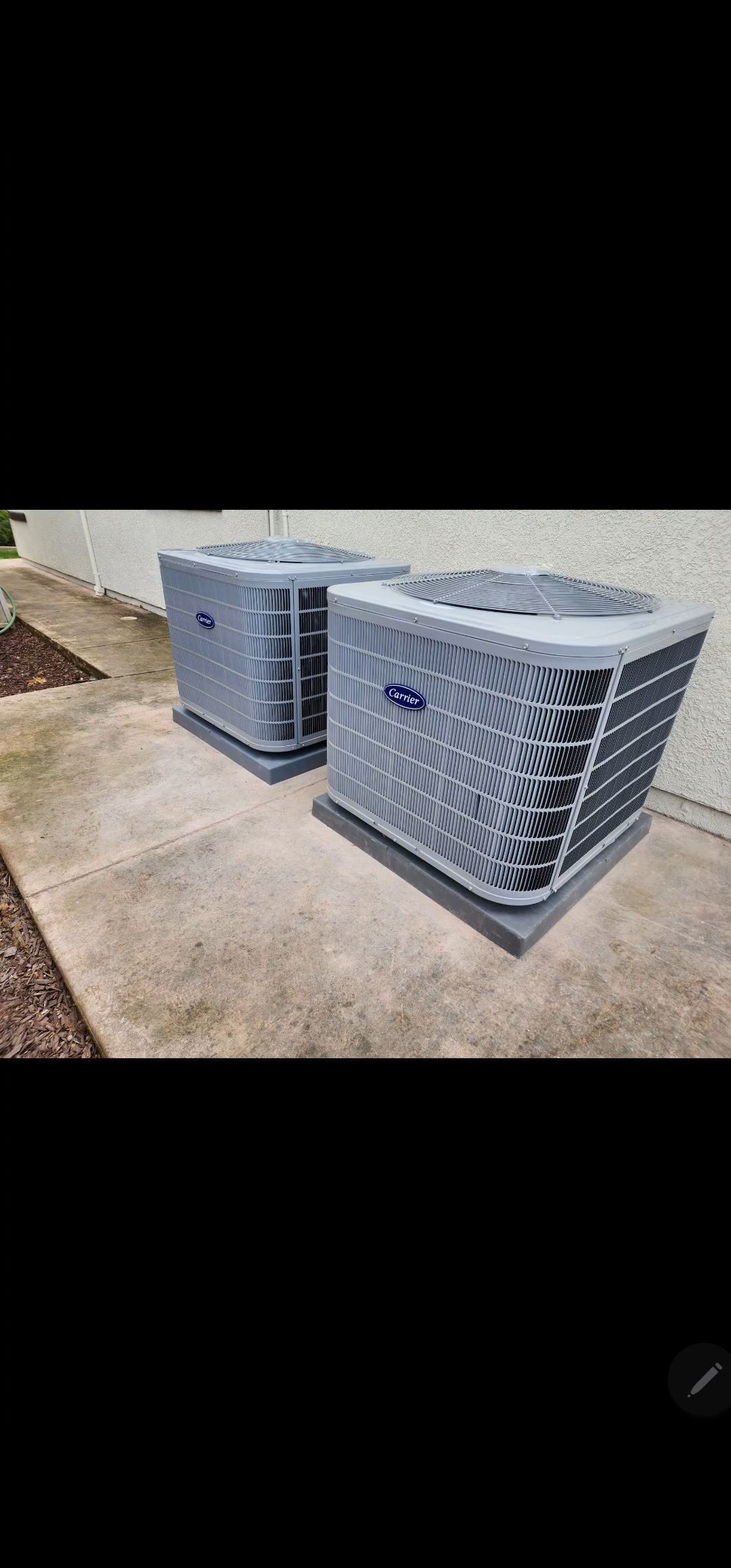 Two Carrier air conditioning units sit on a concrete slab, near a building and mulch.