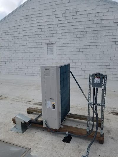 An air conditioning unit on a rooftop, connected to a control box, against a light brick wall.