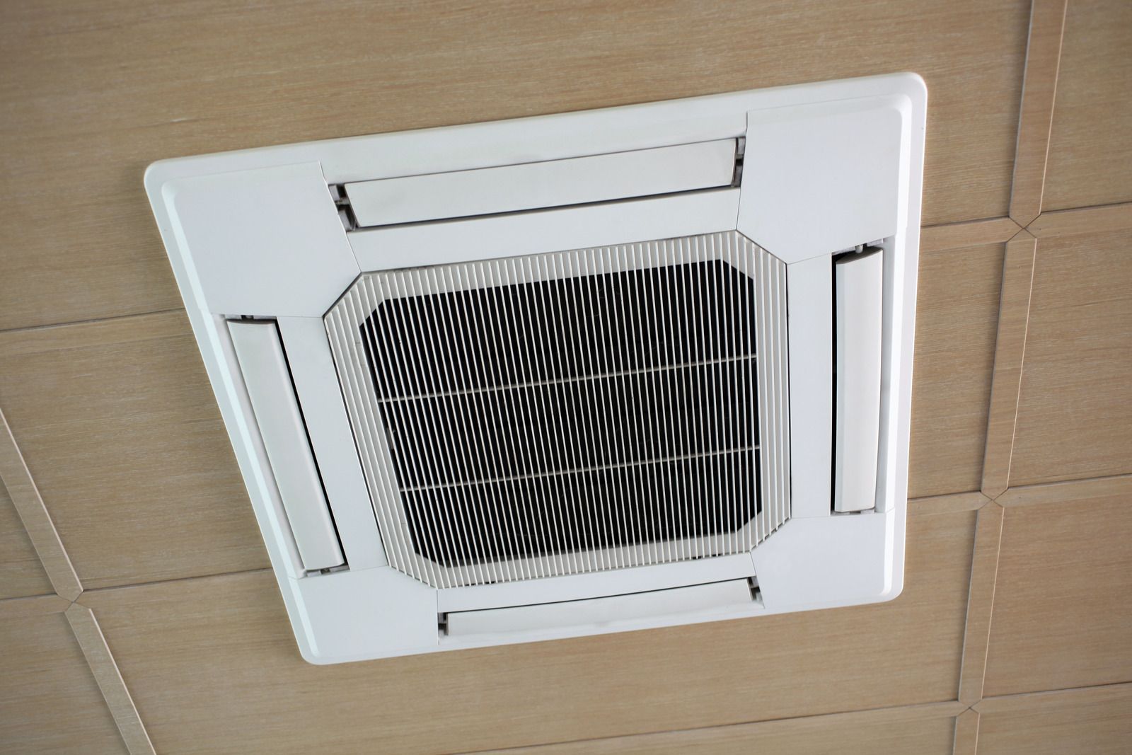 White ceiling-mounted air conditioning unit with black grille, set against a light brown tiled ceiling.