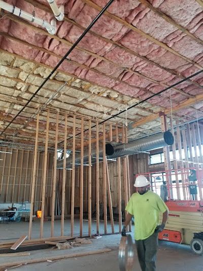 Construction site interior. Man in hard hat, holding metal duct. Wooden studs and exposed ceiling insulation.