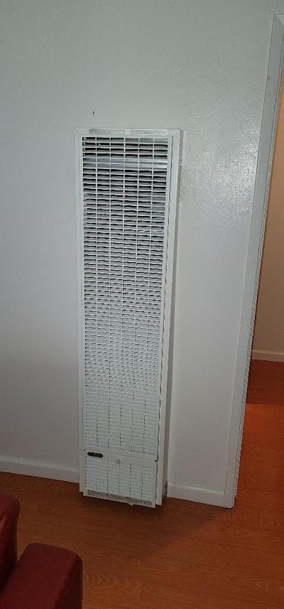 A white wall heater is mounted on a white wall in a room with wood flooring.