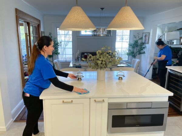 Two people cleaning a kitchen. One wipes a counter, the other sweeps. White cabinets and island.