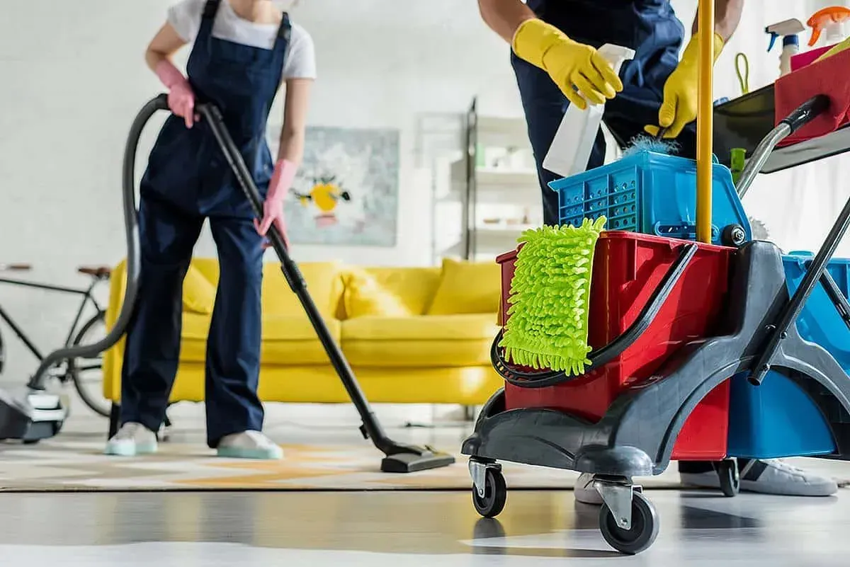 Two people cleaning an apartment; one vacuums, cleaning cart in foreground.