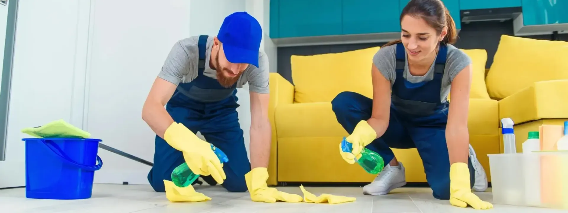 Office cleaners mopping floor, wearing blue overalls and gloves. Caution sign in foreground, desks in background.