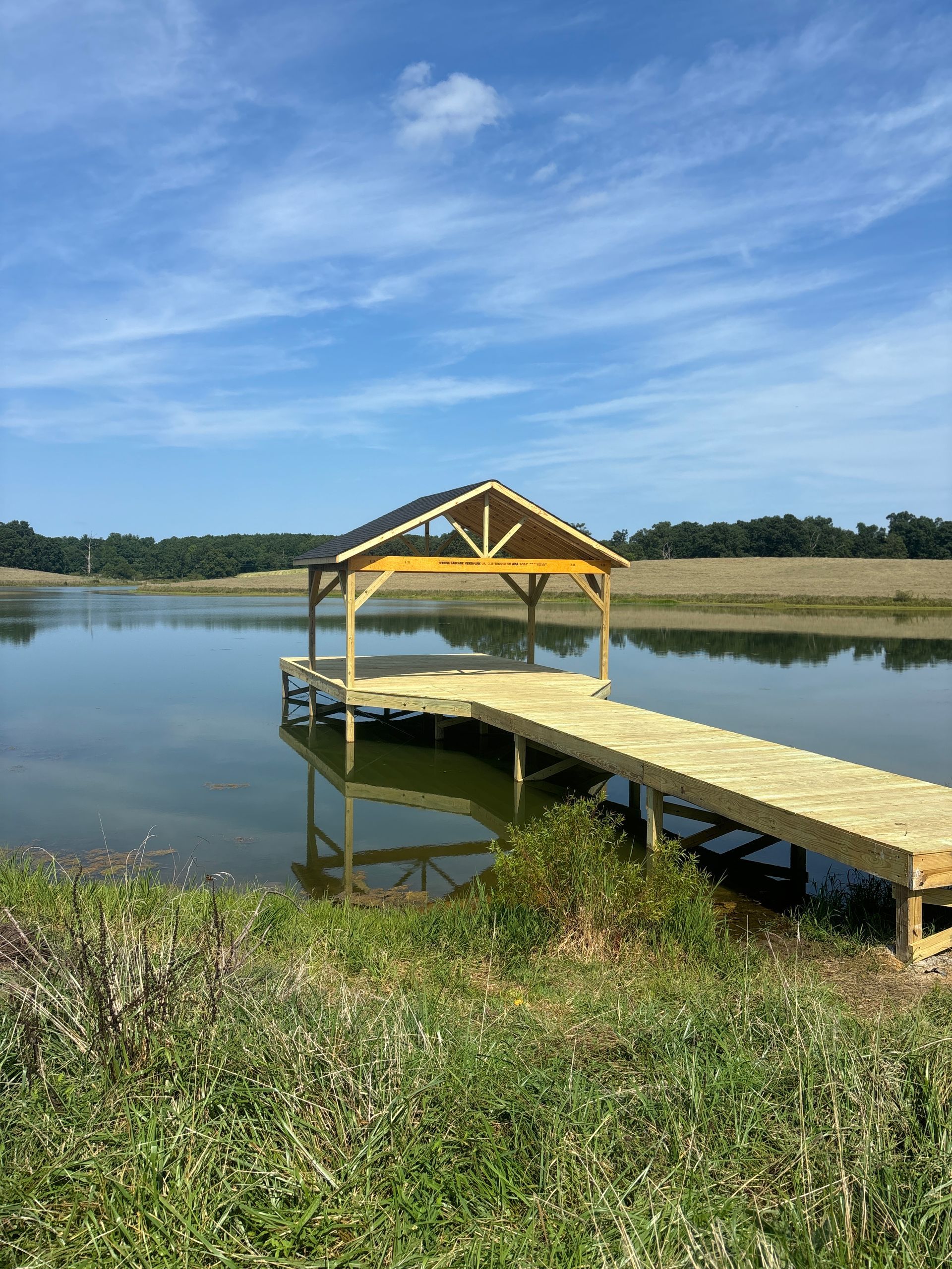 Wooden dock with a covered shelter extends over calm water. Blue sky. Green grass.