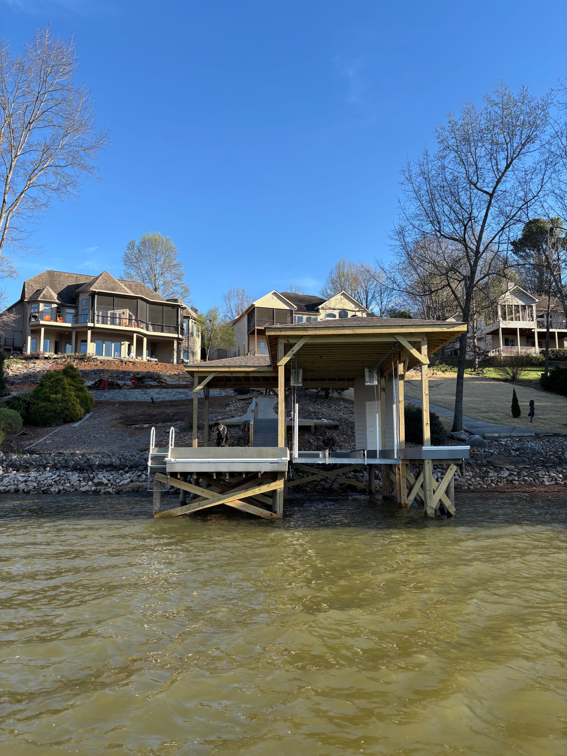A dock on a lake with houses in the background. The water is brown, and the sky is blue.