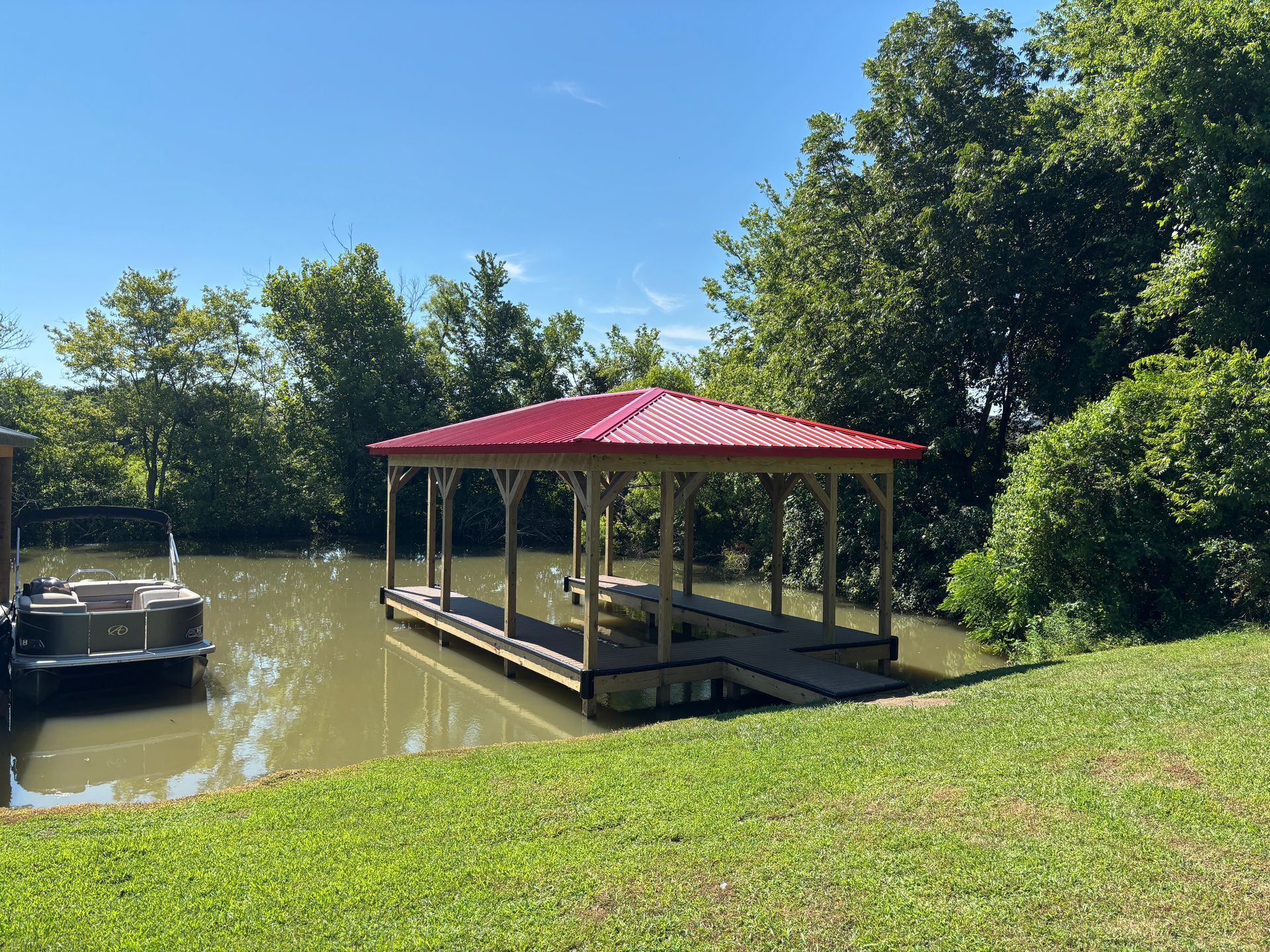 Dock with red roof on calm water, with a boat and green foliage in the background.