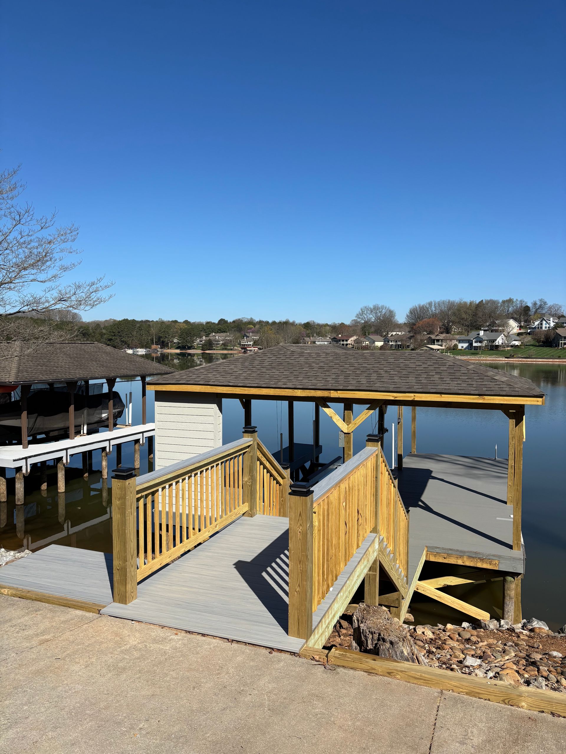 Wooden dock on a lake with a ramp and covered seating area, under a clear blue sky.