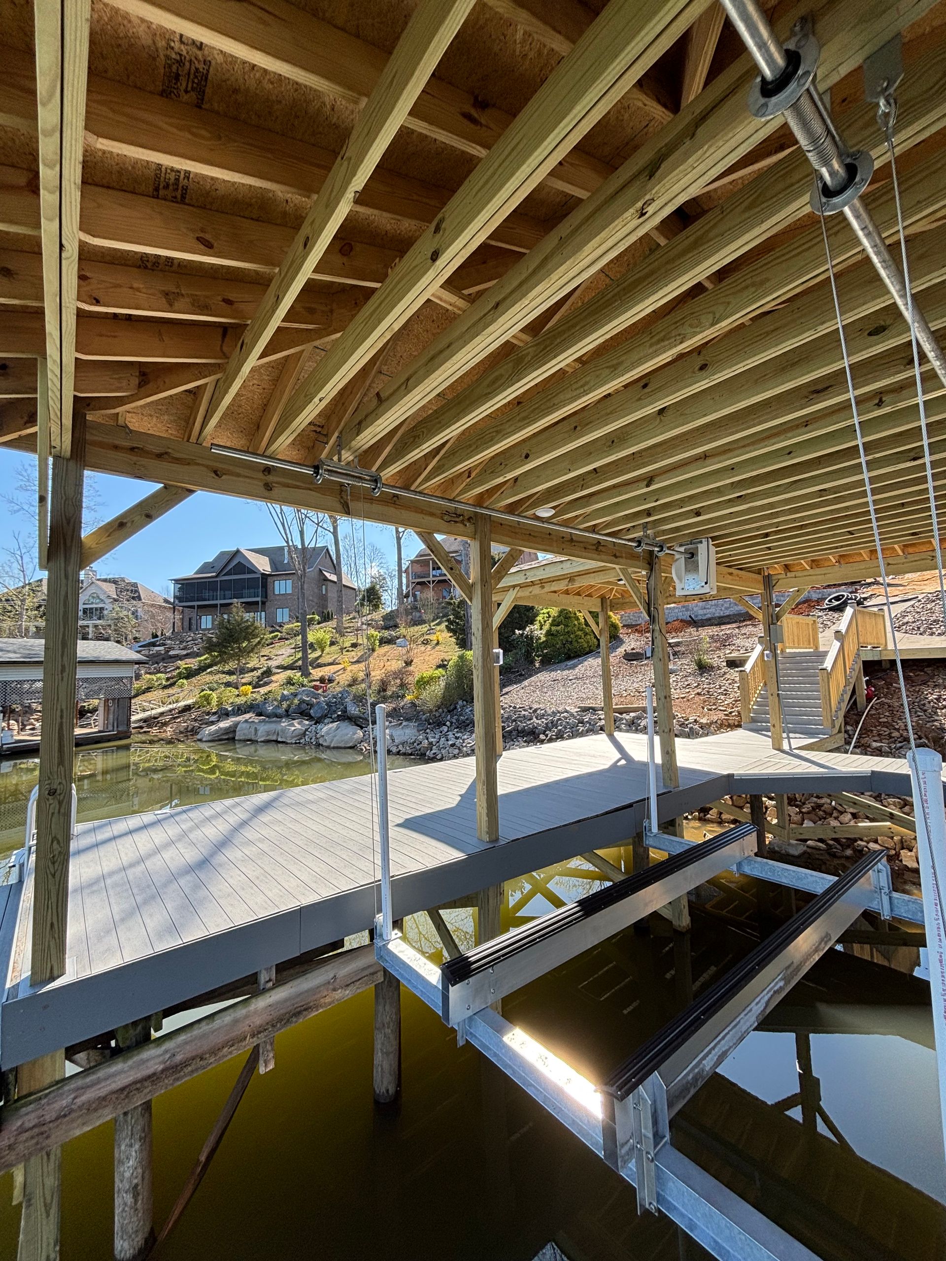 Wooden dock with boat lift over water, under a covered deck. Buildings on a distant hillside.