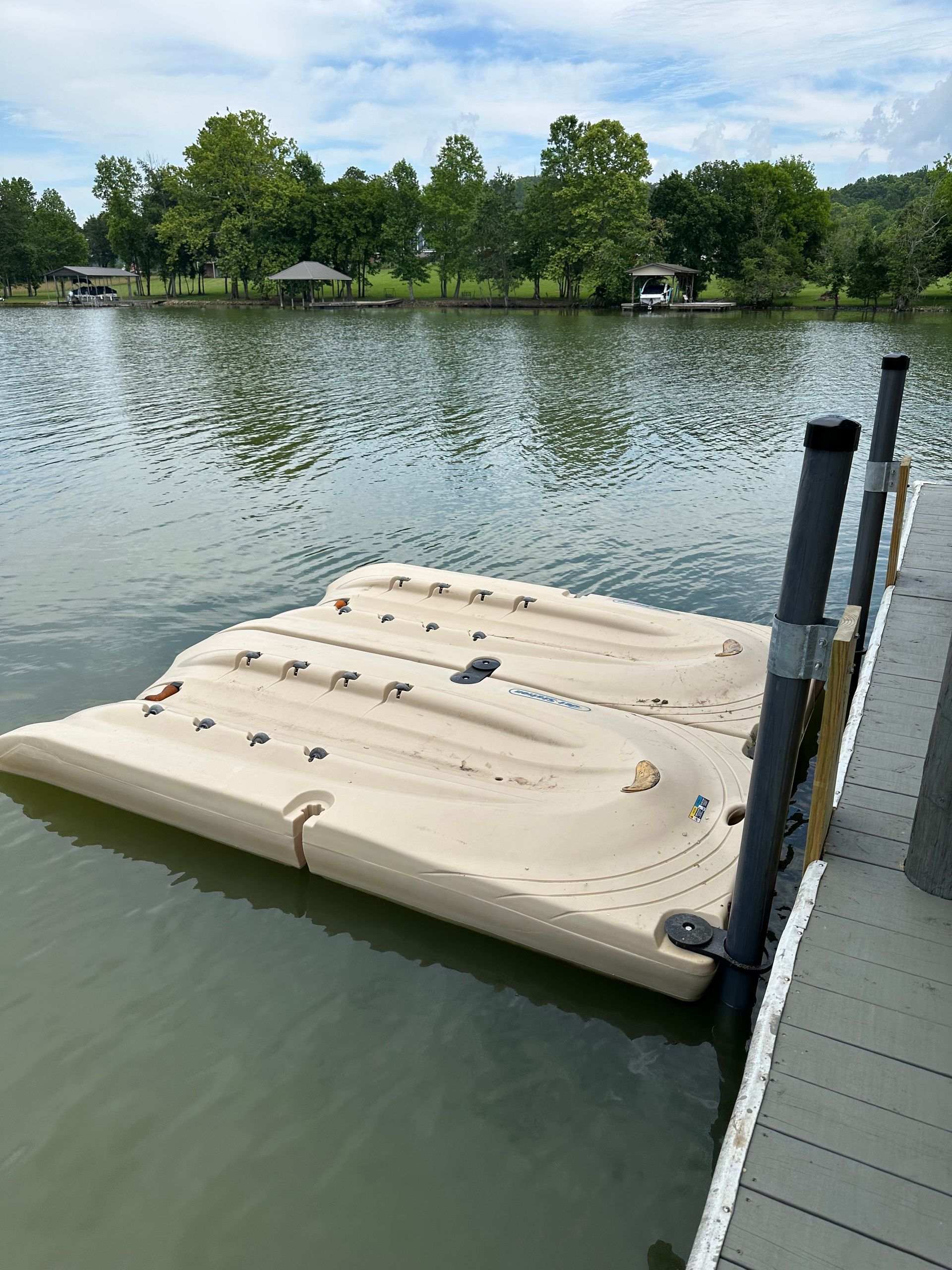 Floating dock on a lake, tan color, attached to a wooden dock with calm water and trees in the background.