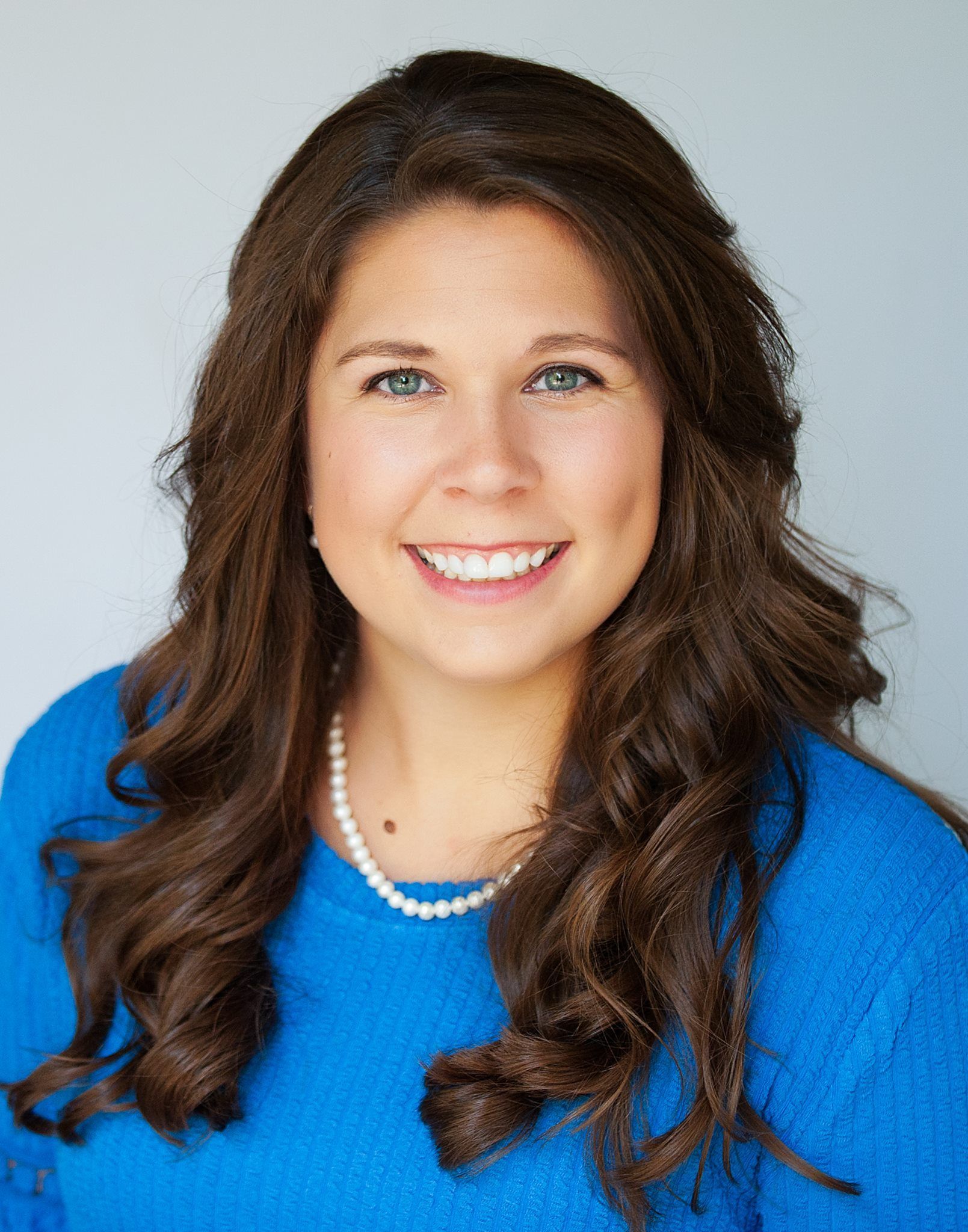 A woman wearing a blue sweater and a pearl necklace is smiling for the camera.