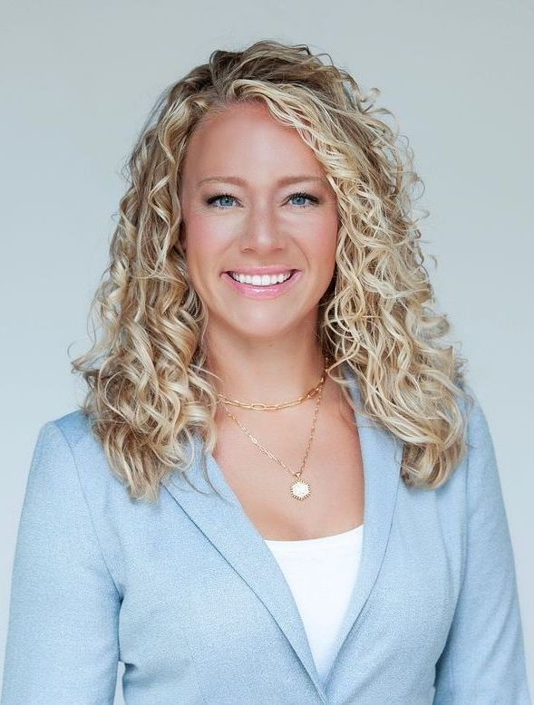 A woman with curly hair is wearing a blue jacket and smiling.