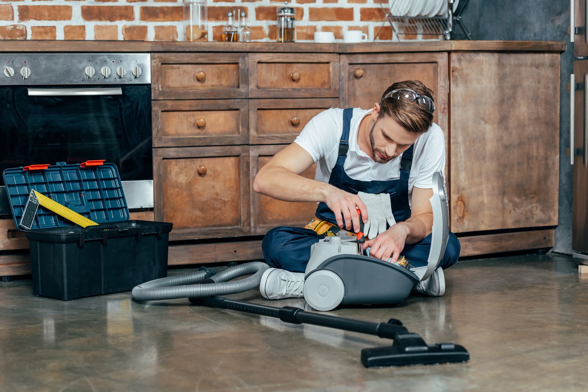 A man is sitting on the floor fixing a vacuum cleaner in a kitchen.