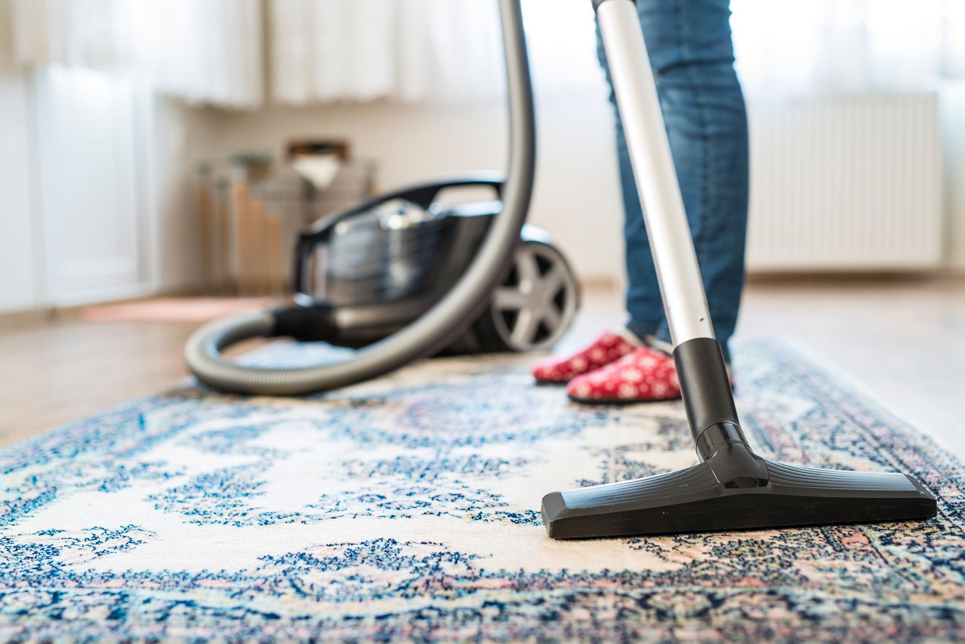 Person vacuuming patterned rug with modern vacuum cleaner in bright living room.