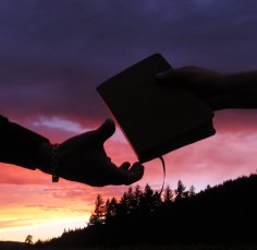 A person is handing another person a bible at sunset