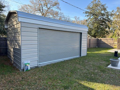 A metal garage with a roll up door is in the backyard of a house.