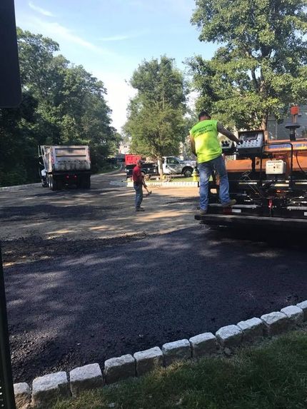 Construction workers operate heavy machinery to pave a driveway with fresh asphalt near a stone-bordered lawn.