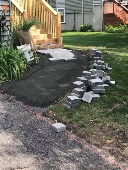 A residential backyard scene shows a paver patio construction project in progress next to wooden stairs and a green lawn.
