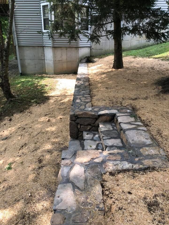 A stone retaining wall and built-in stone steps on a sloping, wood-chipped yard next to a house with gray siding.