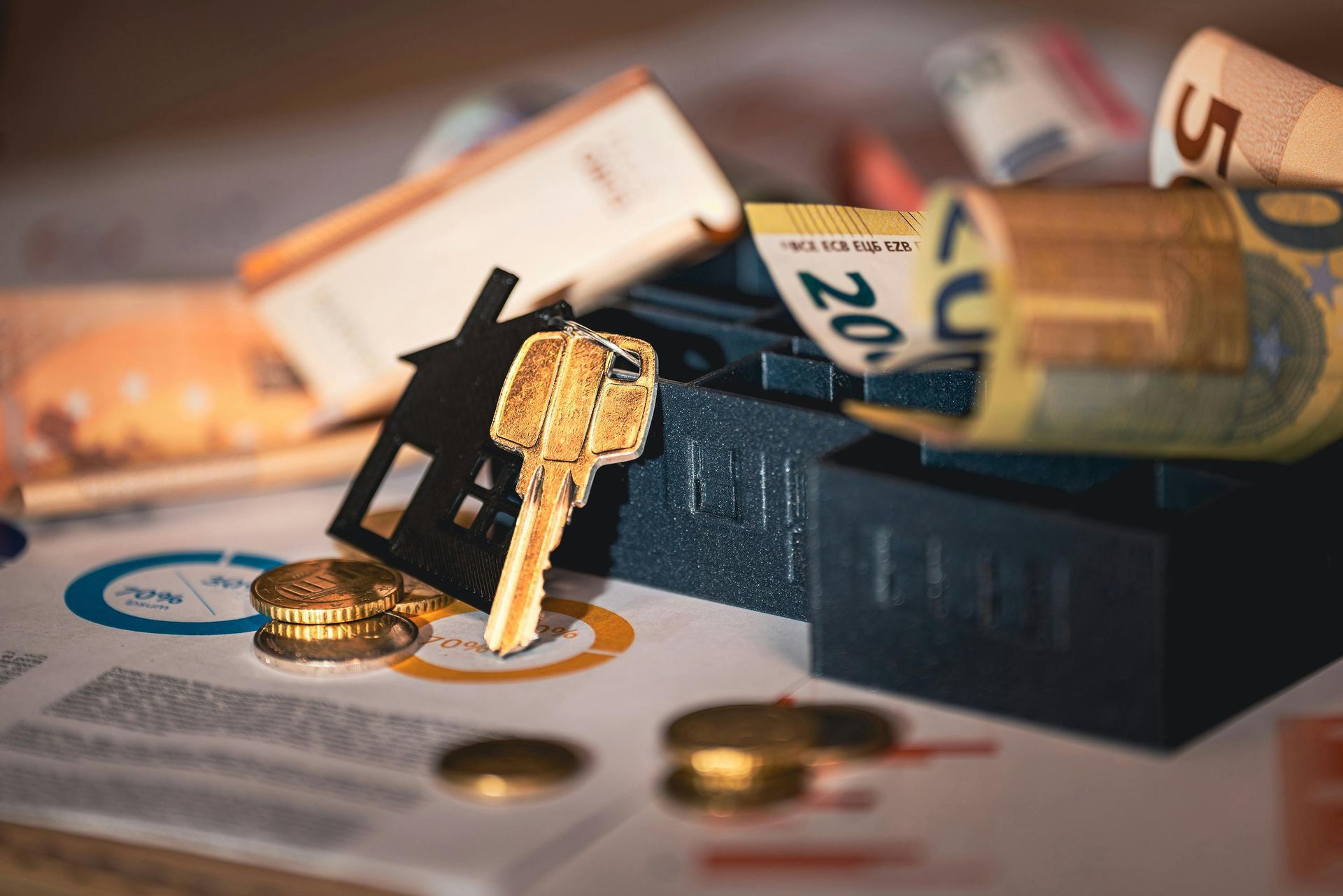 A gold key and a black house silhouette resting on financial charts, surrounded by euro banknotes and coins.