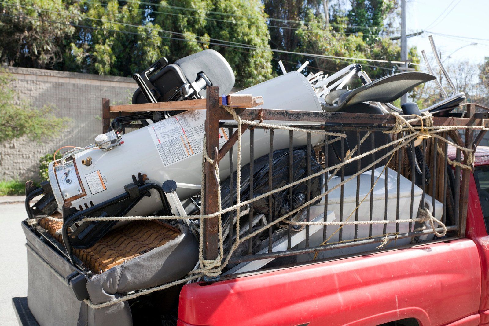 Red truck bed loaded with scrap metal, including a water heater and metal gate, secured with rope.