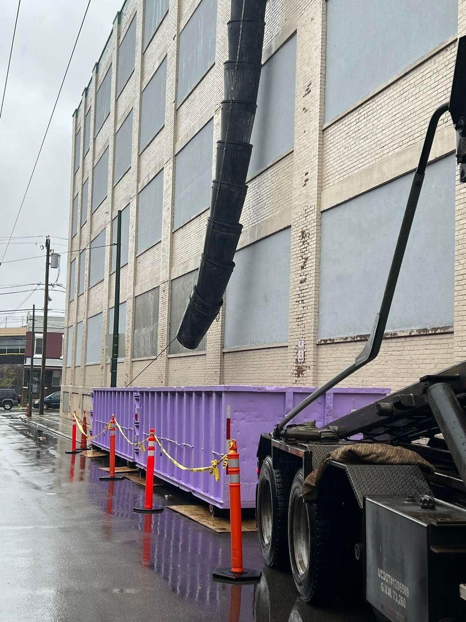 A large purple dumpster next to a building receiving debris via a black chute, on a rainy street.
