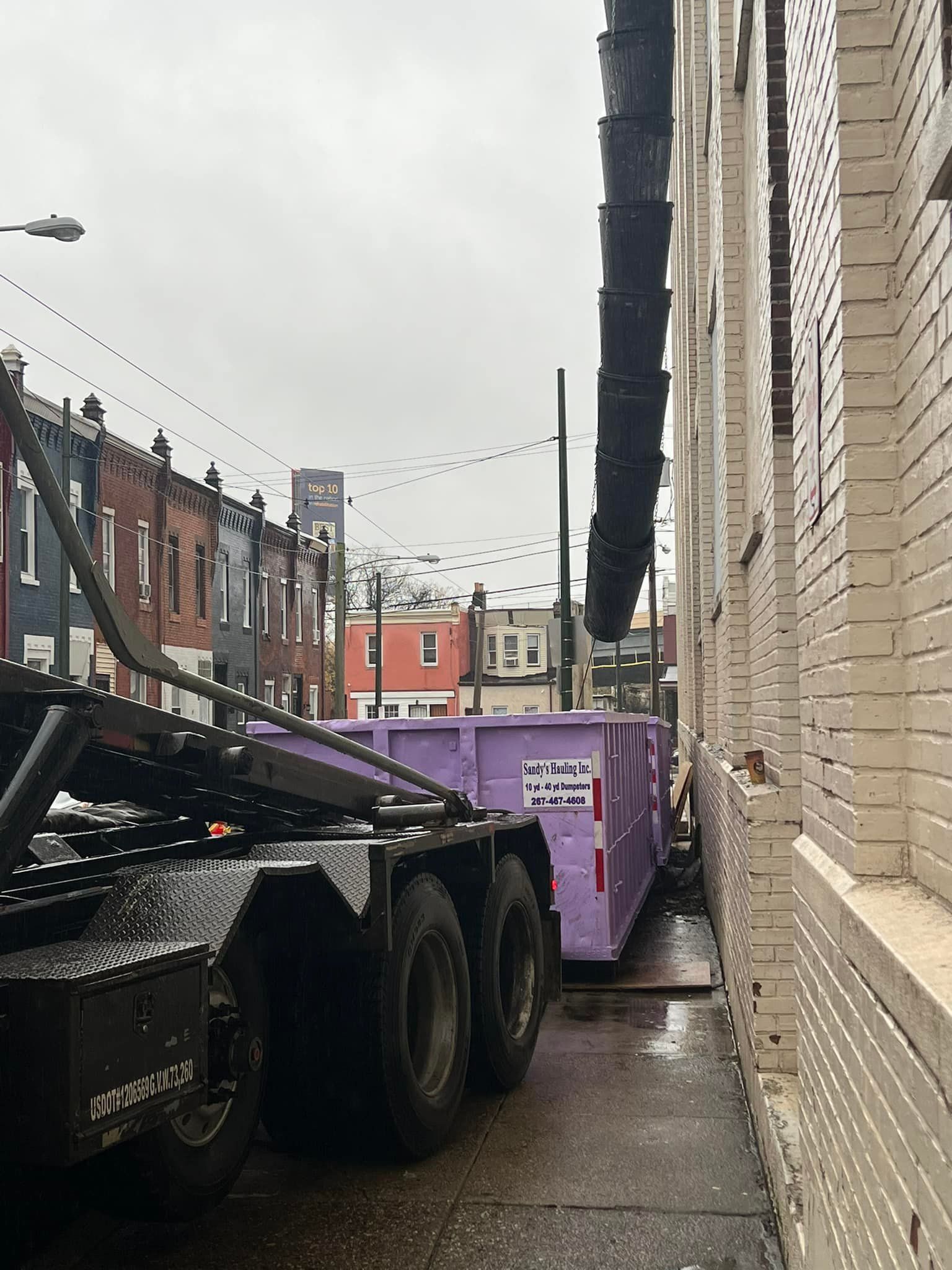 A large truck delivering a purple dumpster in a narrow alley next to a brick building.