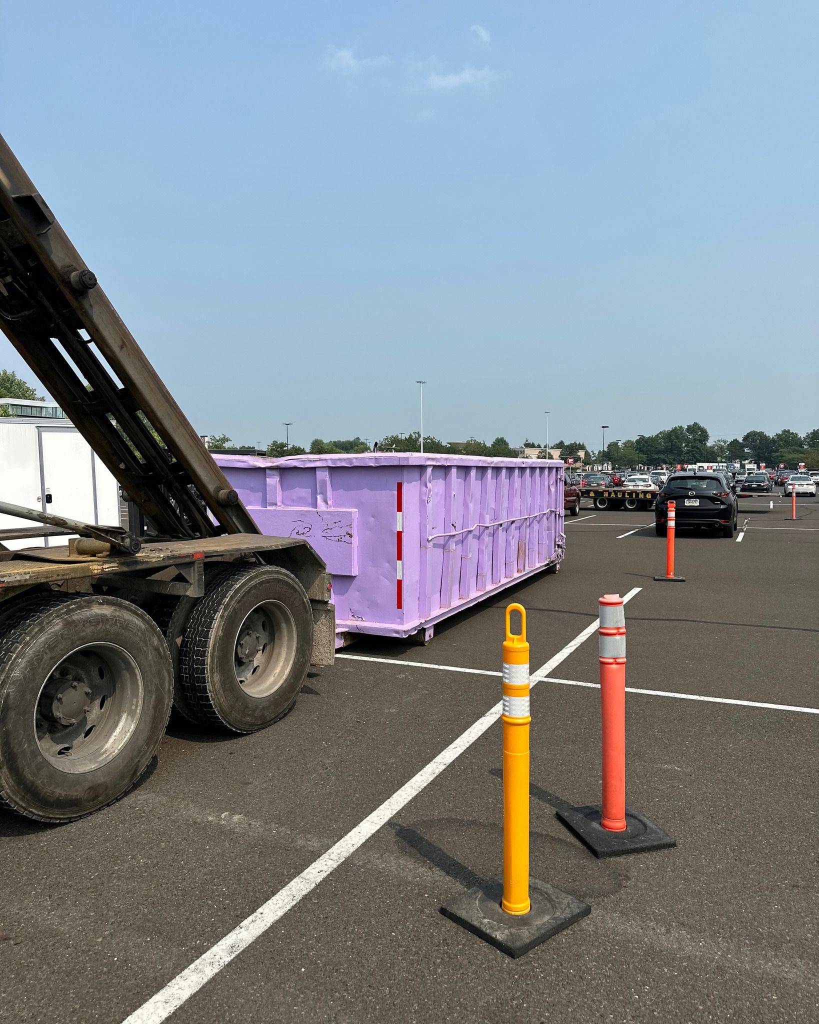 A purple dumpster being unloaded from a truck in a parking lot, next to traffic cones.