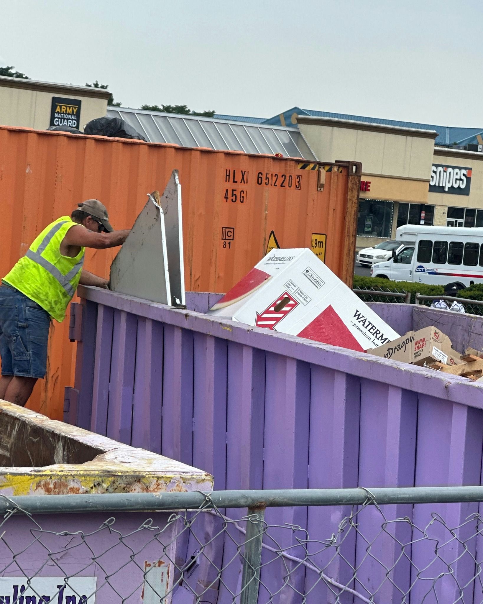 Worker in neon vest tossing debris into a purple dumpster, near a rusty orange container in front of a Staples store.