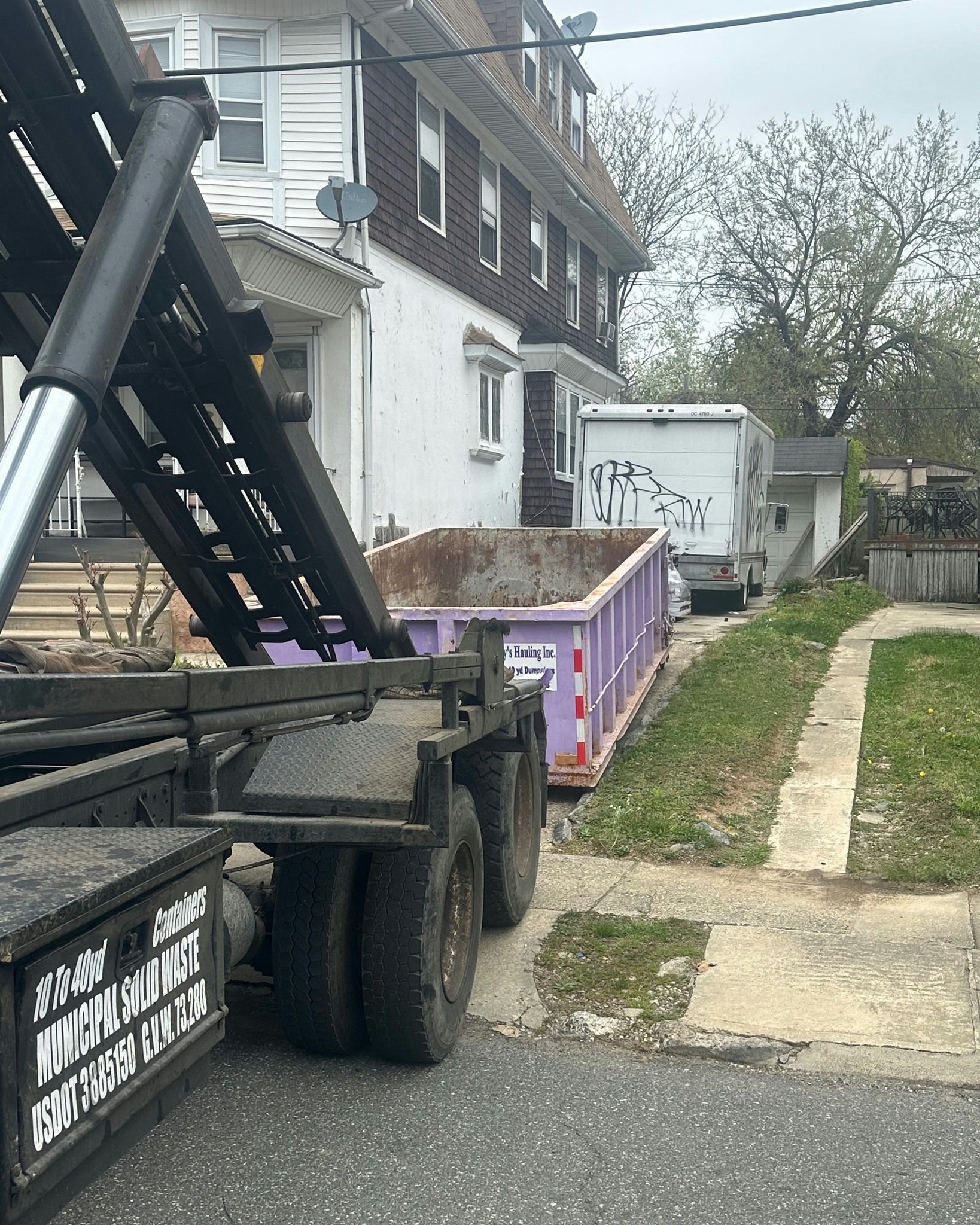 Dump truck with a purple dumpster, parked on a residential street, in front of a multi-story house.