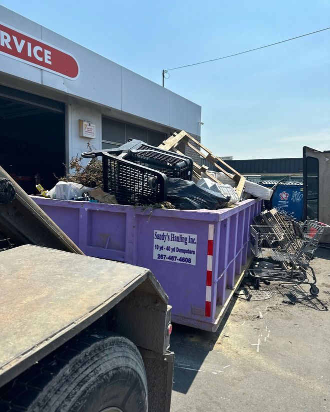 Blue and white dumpster filled with wooden debris, parked on a brick surface near a garage and car.