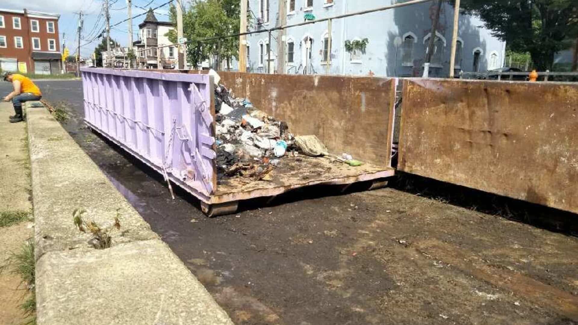 Green dumpster filled with construction debris parked on a driveway with a house and trees in the background.