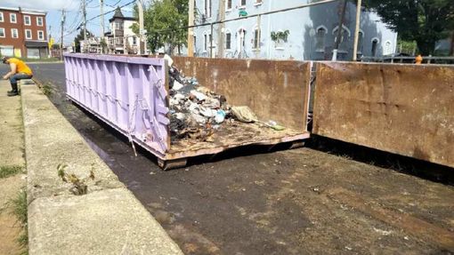 Purple and rust-colored dumpsters overflowing with trash on a street, a person in orange shirt nearby.