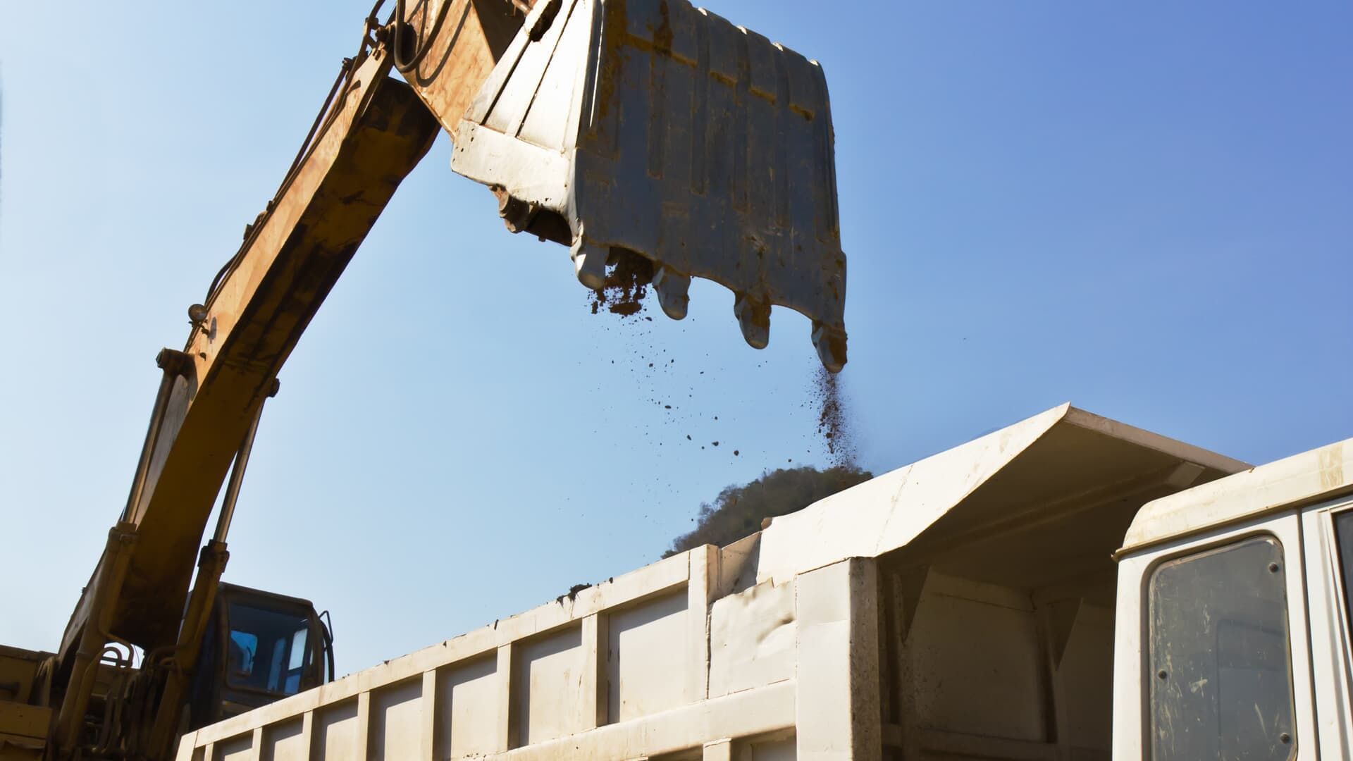 An excavator dumping dirt into the bed of a truck under a blue sky.