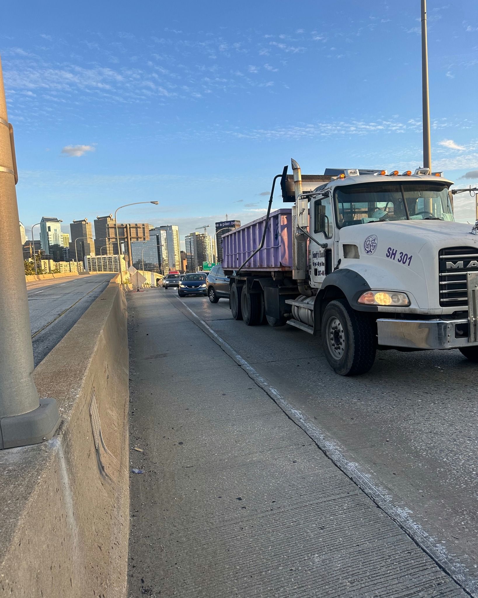 Dump truck on a road, cityscape in the background. Clear sky, concrete barriers, and other vehicles present.